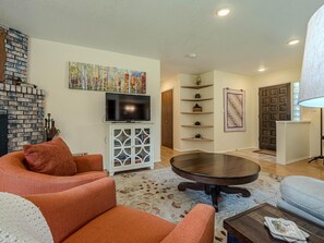 A living room with orange sofas, a TV on a white cabinet, a fireplace, and a door leading to another room. - Tanglewood: Dog-Friendly Retreat + Spacious Yard (Grand Junction)
