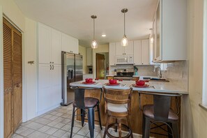 A kitchen with white cabinets, a stainless steel refrigerator, a microwave, and a stove. There are three bar stools and two pendant lights. - Tanglewood: Dog-Friendly Retreat + Spacious Yard (Grand Junction)