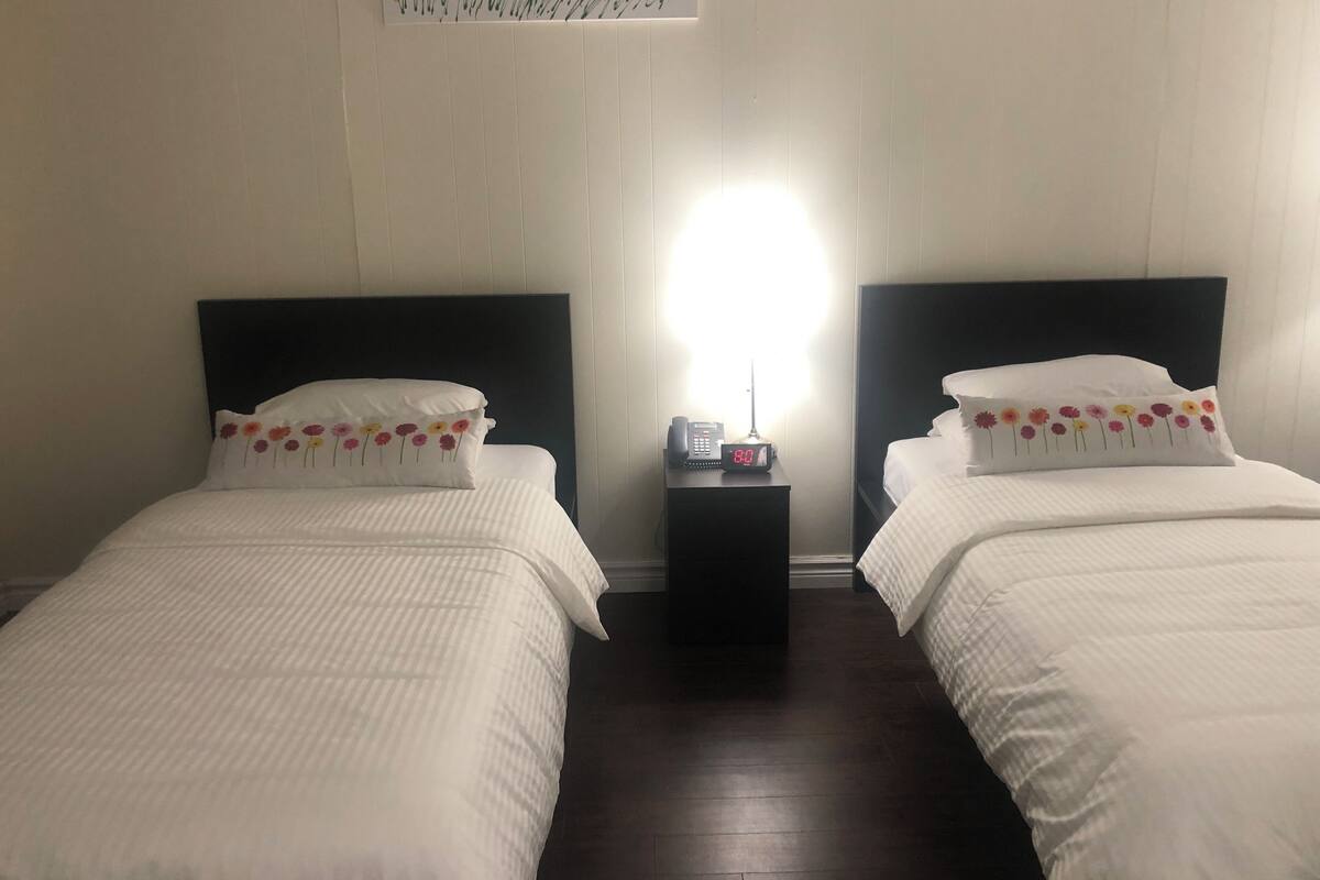 Ein Hotelzimmer mit zwei Einzelbetten, einem Nachttisch mit Telefon und digitaler Uhr sowie einer Wandleuchte.