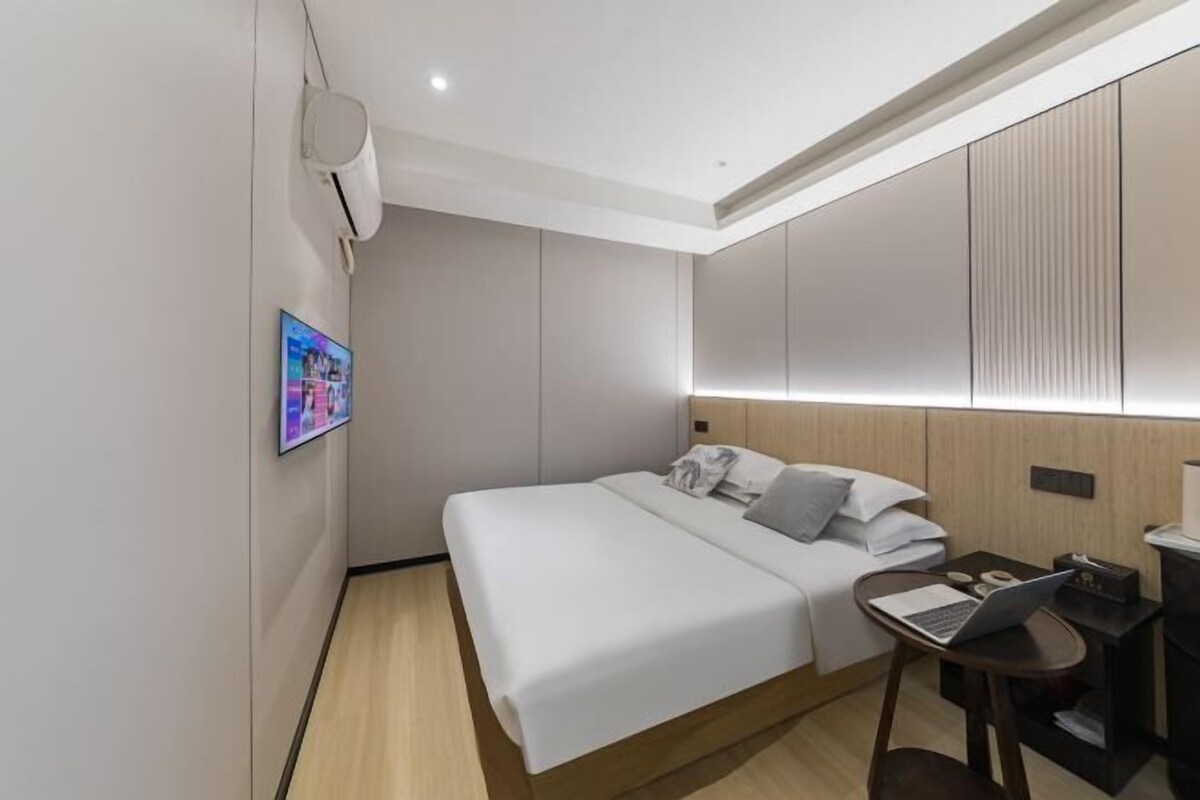 A modern hotel room with a neatly made bed, a laptop on a small table, and a wall-mounted air conditioner.