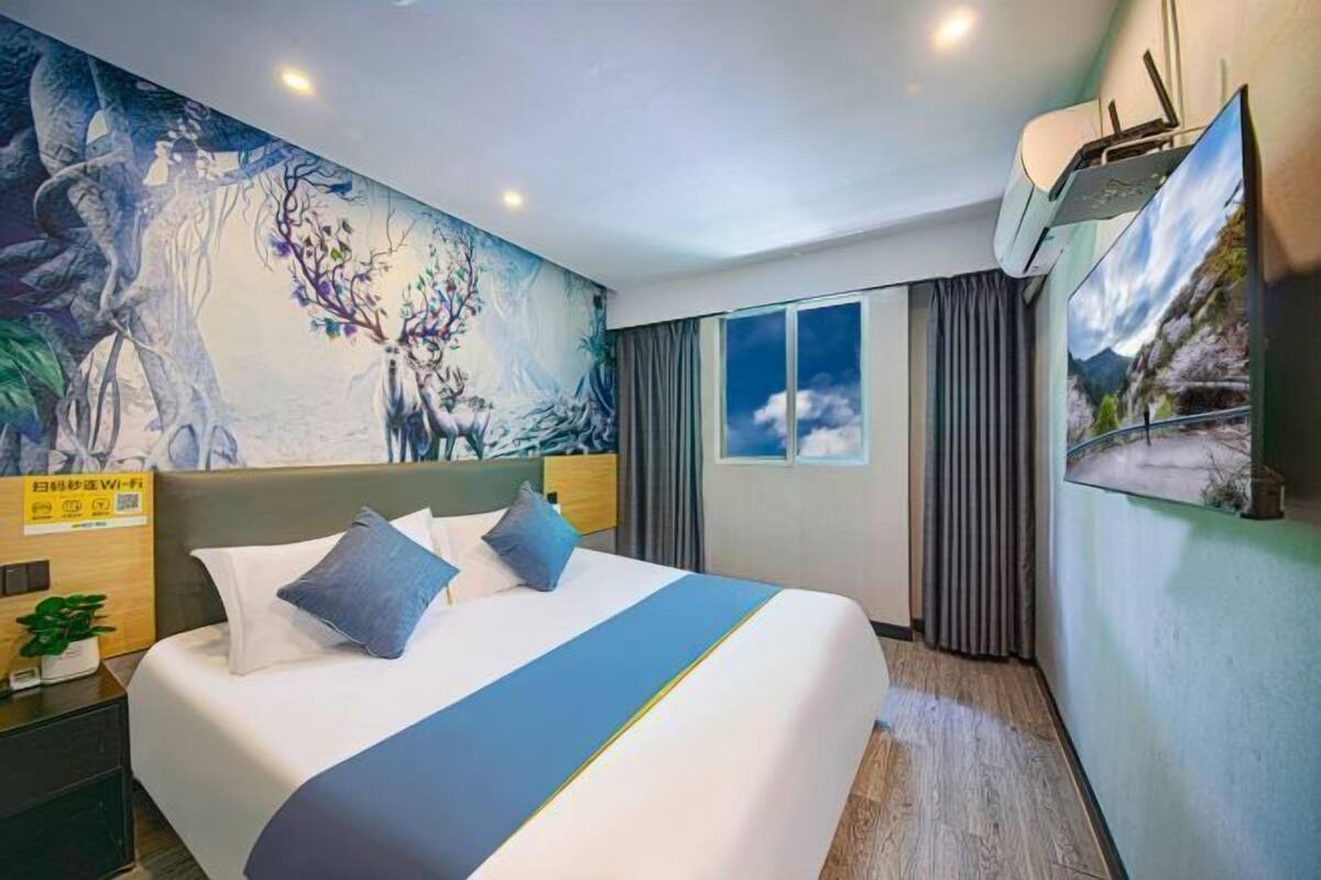 Habitación de hotel con un mural grande en la pared, una cama con ropa de cama azul, una ventana con cortinas y un aire acondicionado instalado.