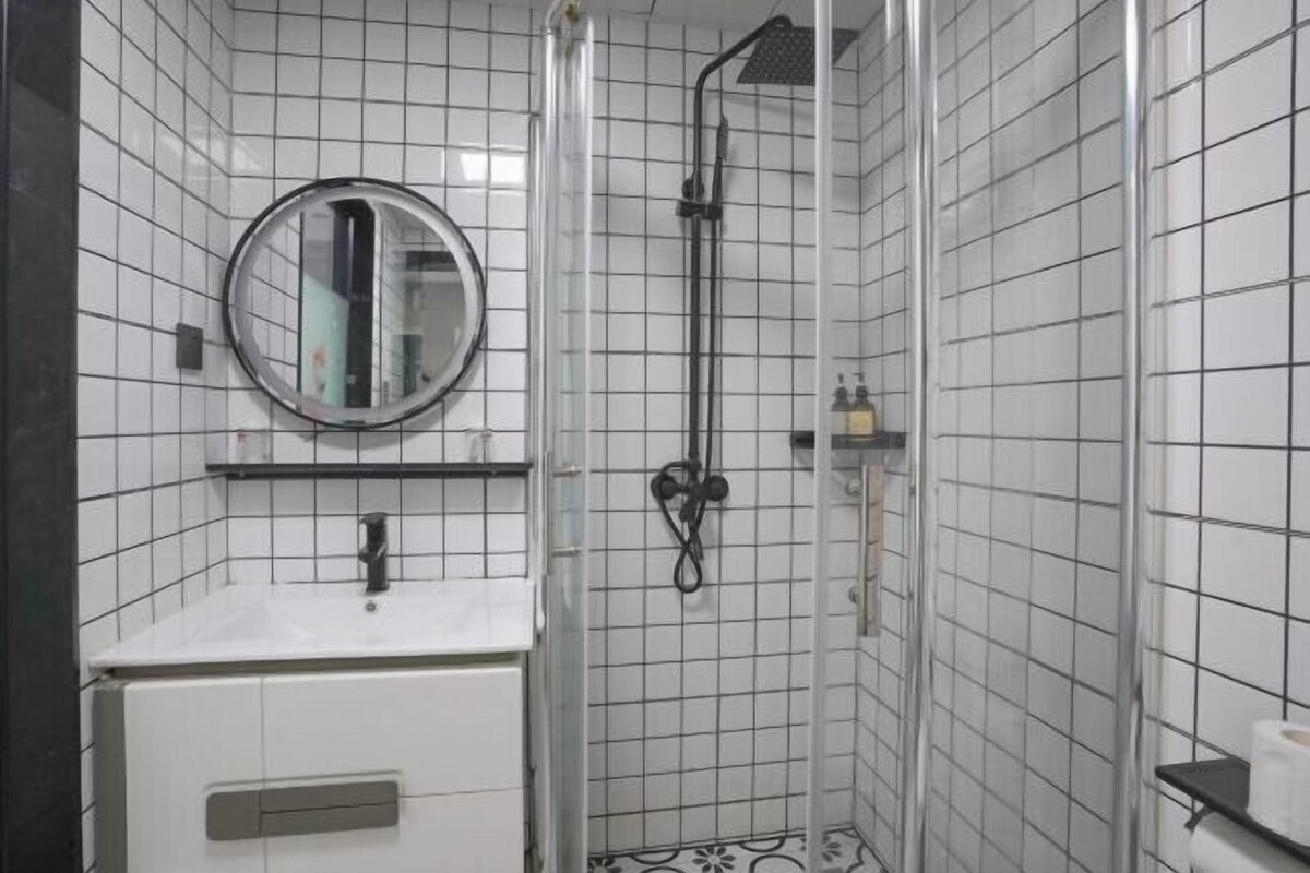 A bathroom with white tiled walls, a round mirror, a sink, and a shower enclosure.