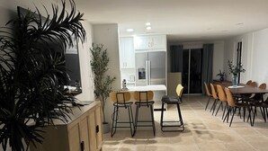 Eine moderne Küche mit weißen Schränken, einem Einbauofen und einem Kühlschrank. Es gibt einen Essbereich mit einem Holztisch und Stühlen, sowie eine große Küsterpflanze.