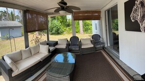 Eine überdachte Terrasse mit Korbmöbeln, einem Glastisch und einem Deckenventilator.