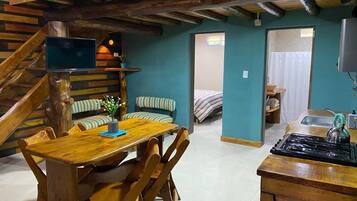 Un interior rústico con una mesa de comedor de madera, sillas a rayas, un televisor y una zona de cocina con estufa y tetera.