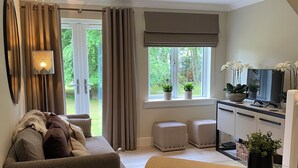 Een moderne woonkamer met een bank, voetenbankjes, een kleine tafel, en een keukenhoek met een televisie en planten.