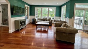 A spacious living room with a large window, a fireplace, and a television mounted on the wall. - NORTH FORK DREAM HOME, 4,000 Sq Ft, 1 Acre, Heated Pool! (Southold)