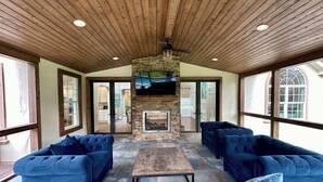 A covered patio area with a stone fireplace, blue sofas, and a television mounted on the wall. - NORTH FORK DREAM HOME, 4,000 Sq Ft, 1 Acre, Heated Pool! (Southold)