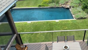 A deck with a pool, wooden chairs, and a table with a bowl of fruit. - NORTH FORK DREAM HOME, 4,000 Sq Ft, 1 Acre, Heated Pool! (Southold)