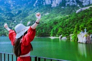 Una persona con remera roja y sombrero blanco de pie junto a un lago, con vegetación y rocas al fondo.