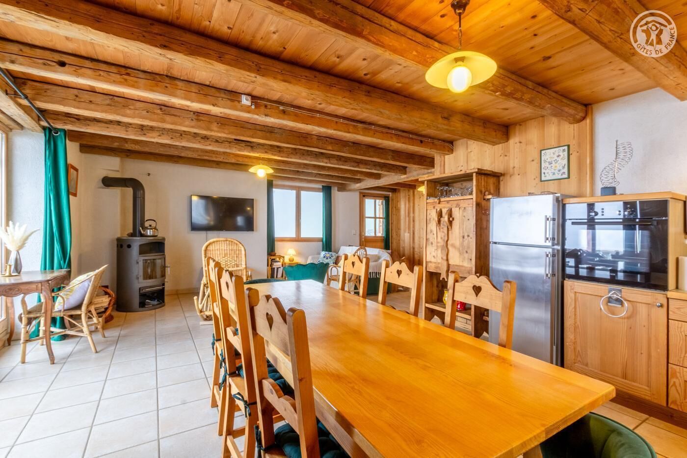 Un intérieur rustique en bois, avec une longue table à manger, des chaises en bois, un poêle encastré, un téléviseur à écran plat et une cuisine moderne équipée d’un réfrigérateur et d’un four.