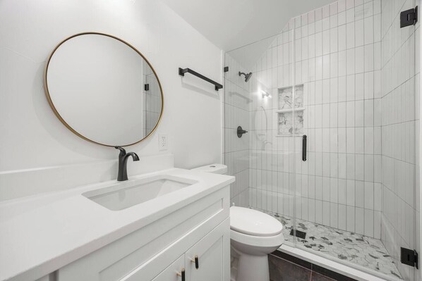 A modern bathroom with a white vanity, a round mirror with a gold frame, a toilet, and a shower area with tiled walls.