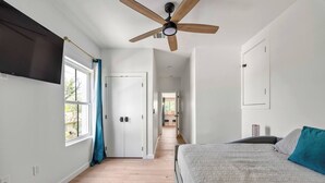 Ein Schlafzimmer mit einem Bett, einem Deckenventilator, einem Fenster mit Vorhängen und einem Flur, der in einen anderen Raum führt.