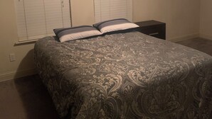 Une chambre à coucher avec un grand lit, deux tables de chevet et deux fenêtres équipées de stores.