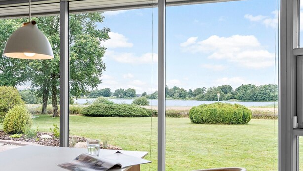 A dining area with a table, chairs, and a hanging light fixture. A window offers a view of a grassy area with trees and a body of water. - Lovely home in Grindsted with lake view (Grindsted)