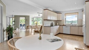 A modern kitchen with white cabinets, a dining area with a round table, and a view of greenery outside. - Lovely home in Grindsted with lake view (Grindsted)