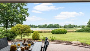 An outdoor patio with a table set for breakfast, overlooking a lake and a well-maintained lawn. - Lovely home in Grindsted with lake view (Grindsted)