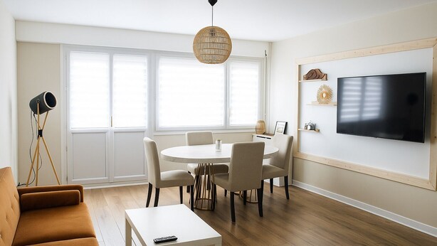 A modern living room with a round dining table, beige sofas, a flat-screen TV, and a large window with blinds.
