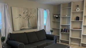 Ein Wohnzimmer mit schwarzem Sofa, passendem Hocker, Bücherregal und einem Bild an der Wand.