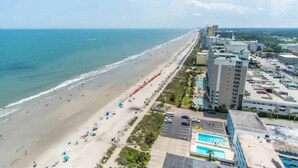 An aerial view of a beach with a row of beach umbrellas, a swimming pool, and adjacent buildings.