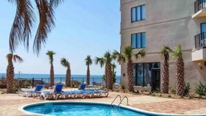 A swimming pool with lounge chairs and palm trees, adjacent to a building with large windows and a view of the sea.