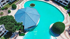 An aerial view of a swimming pool area with a large blue umbrella, surrounded by well-maintained greenery and modern buildings. - Riviera del Mar (Puerto Plata)