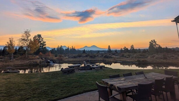A patio with a table and chairs overlooking a lake with a waterfall, surrounded by trees and a mountain in the background. - Stunning mountain views, quiet, peaceful, unique, private attached guest suite (Bend)