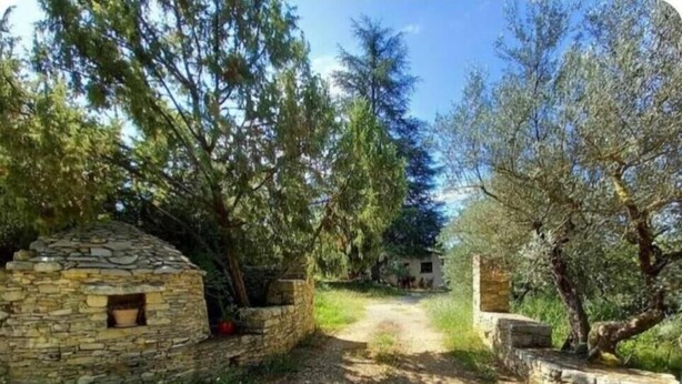 Un edificio de piedra con una ventana pequeña, rodeado de árboles y un sendero de tierra que lleva a una casa.
