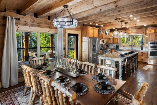A rustic wooden cabin with a large dining table set for six, a kitchen with modern appliances, and large windows offering a view of the forest. - Smoky Mtn Luxury: Views, Theater, Outdoor Kitchen (Sevierville)