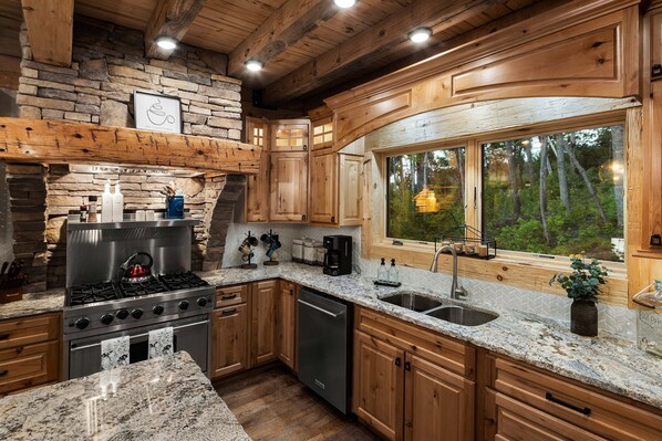 Una cocina moderna con encimeras de granito, armarios de madera, estufa integrada y un amplio ventanal con vistas al bosque.