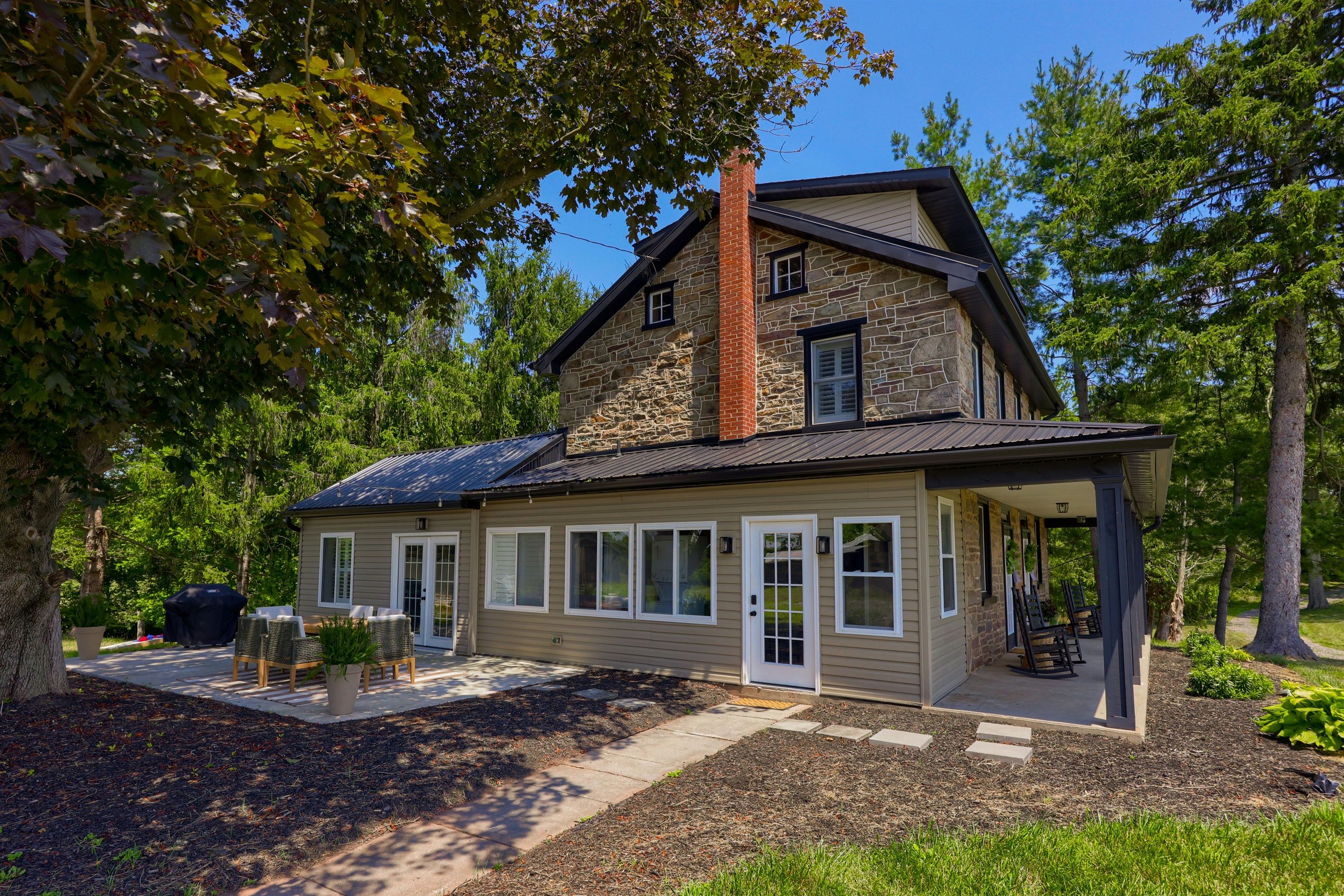 Une maison à deux étages avec une façade en pierre, un porche couvert et un patio aménagé avec du mobilier d’extérieur.