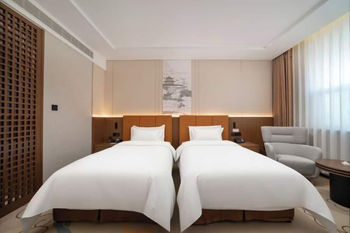 A hotel room with two beds, a wooden headboard, a wall-mounted artwork, a chair, and a small table.