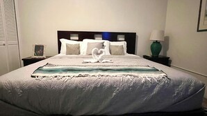 Una cama bien hecha con un arreglo de almohadas en forma de corazón, una mesita de noche con una lámpara y un cuadro en la pared.