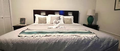 A neatly made bed with a heart-shaped pillow arrangement, a bedside table with a lamp, and a framed picture on the wall.