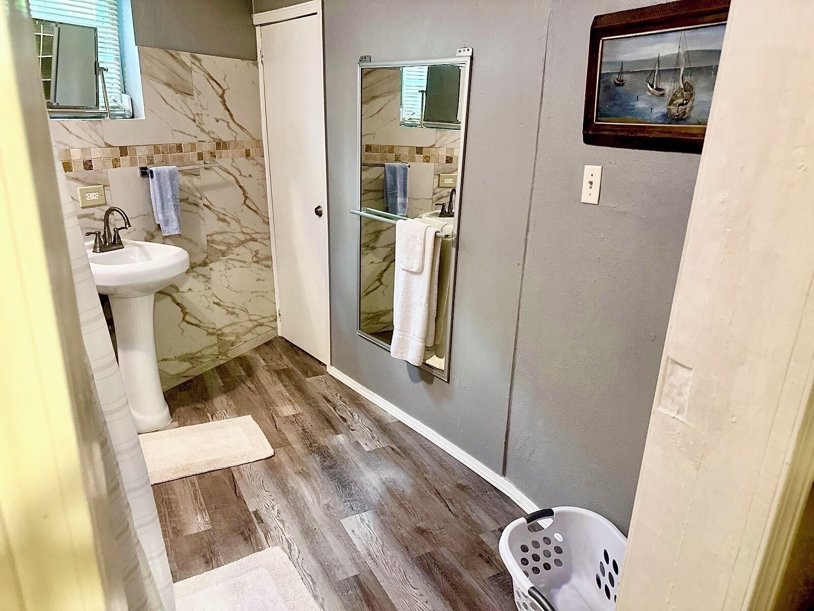 Un baño con pared de azulejos de mármol, un lavabo blanco, mampara de ducha de vidrio y suelo de madera.