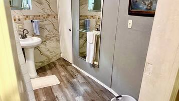 Un baño con pared de azulejos de mármol, un lavabo blanco, mampara de ducha de vidrio y suelo de madera.