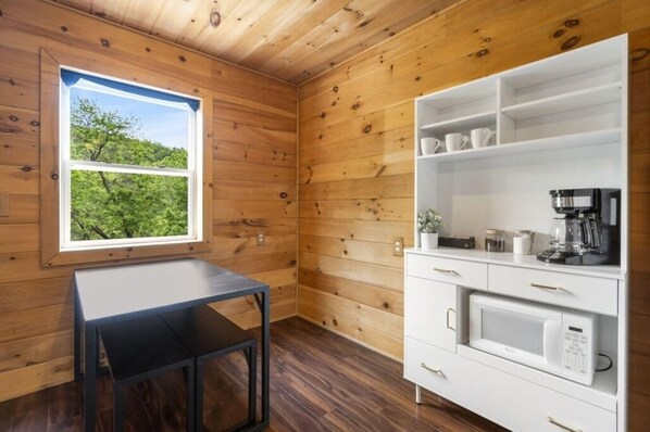 Una habitación con paneles de madera, una ventana, un armario blanco con electrodomésticos de cocina y una mesita.