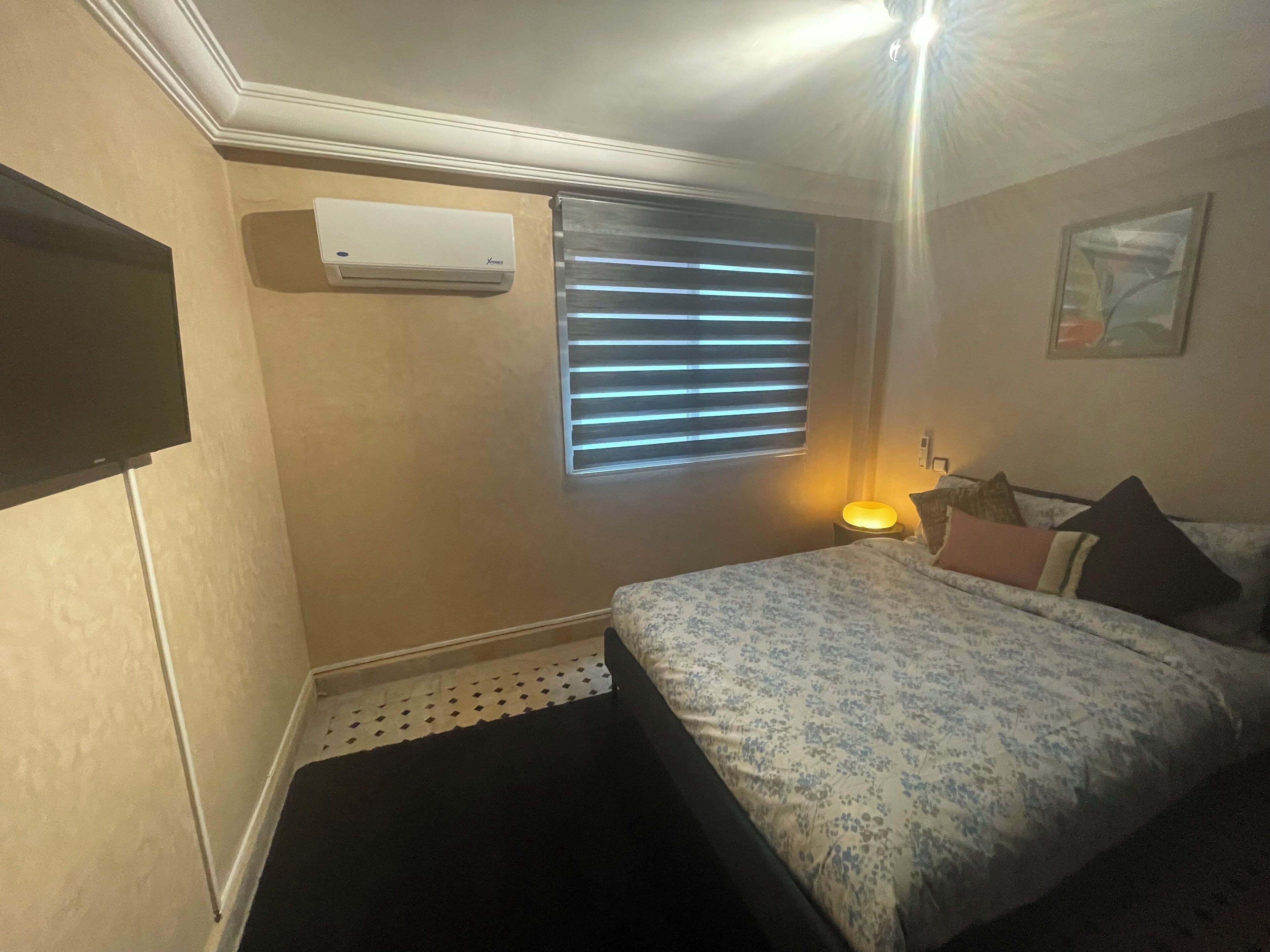 Une chambre à coucher avec un lit, une fenêtre avec des stores, un climatiseur fixé au mur et une petite table de chevet avec une lampe.