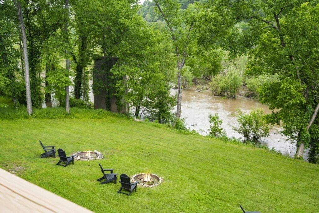 A grassy outdoor area with a fire pit and chairs, overlooking a river.