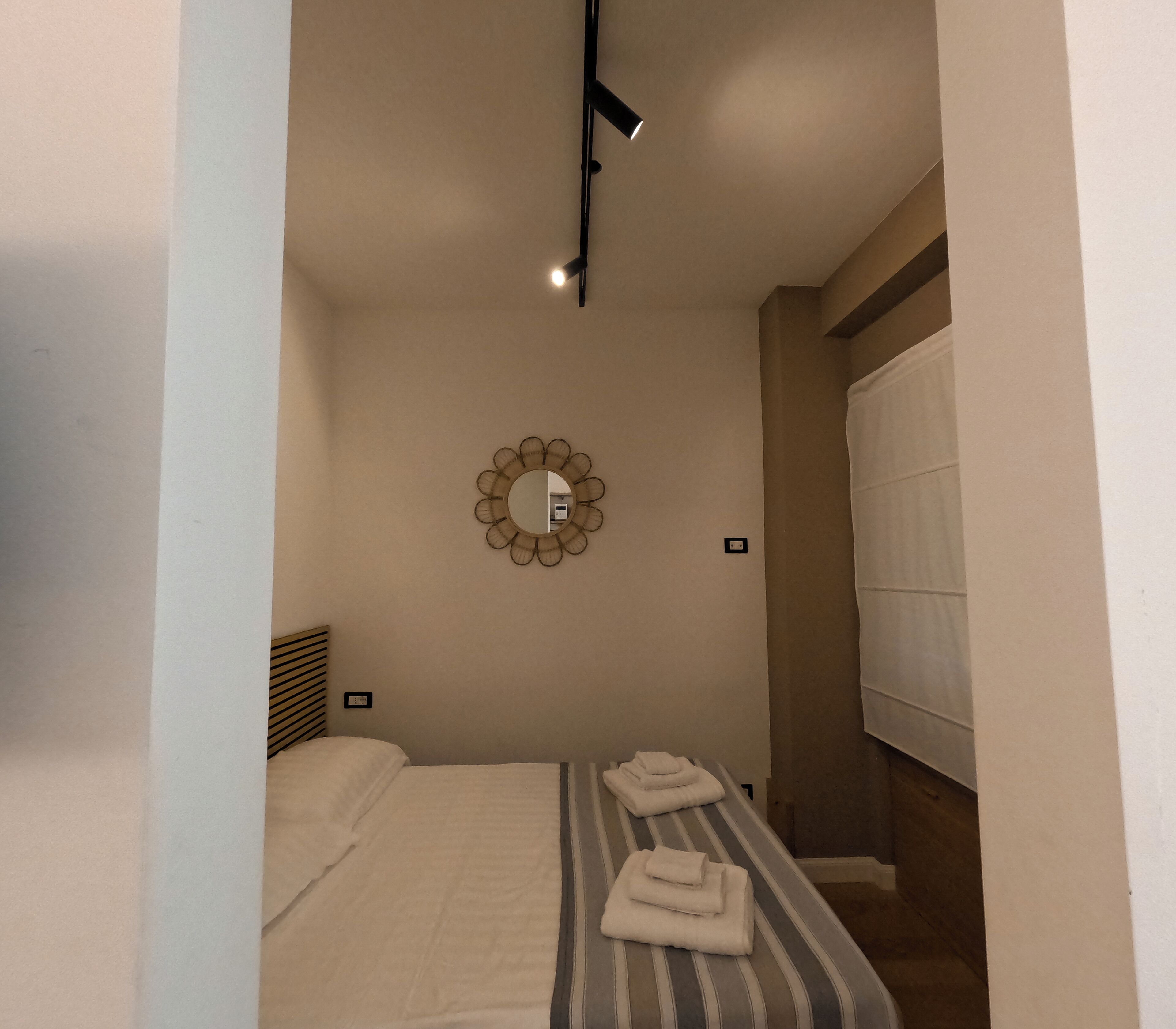 A small, well-lit bedroom with a bed, a bedside table, a mirror, and a window with curtains.