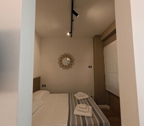 A small, well-lit bedroom with a bed, a bedside table, a mirror, and a window with curtains. - Delizioso Appartamento Metro A Appia Nuova Colli Albani Parco Della Caffarella (Roma)