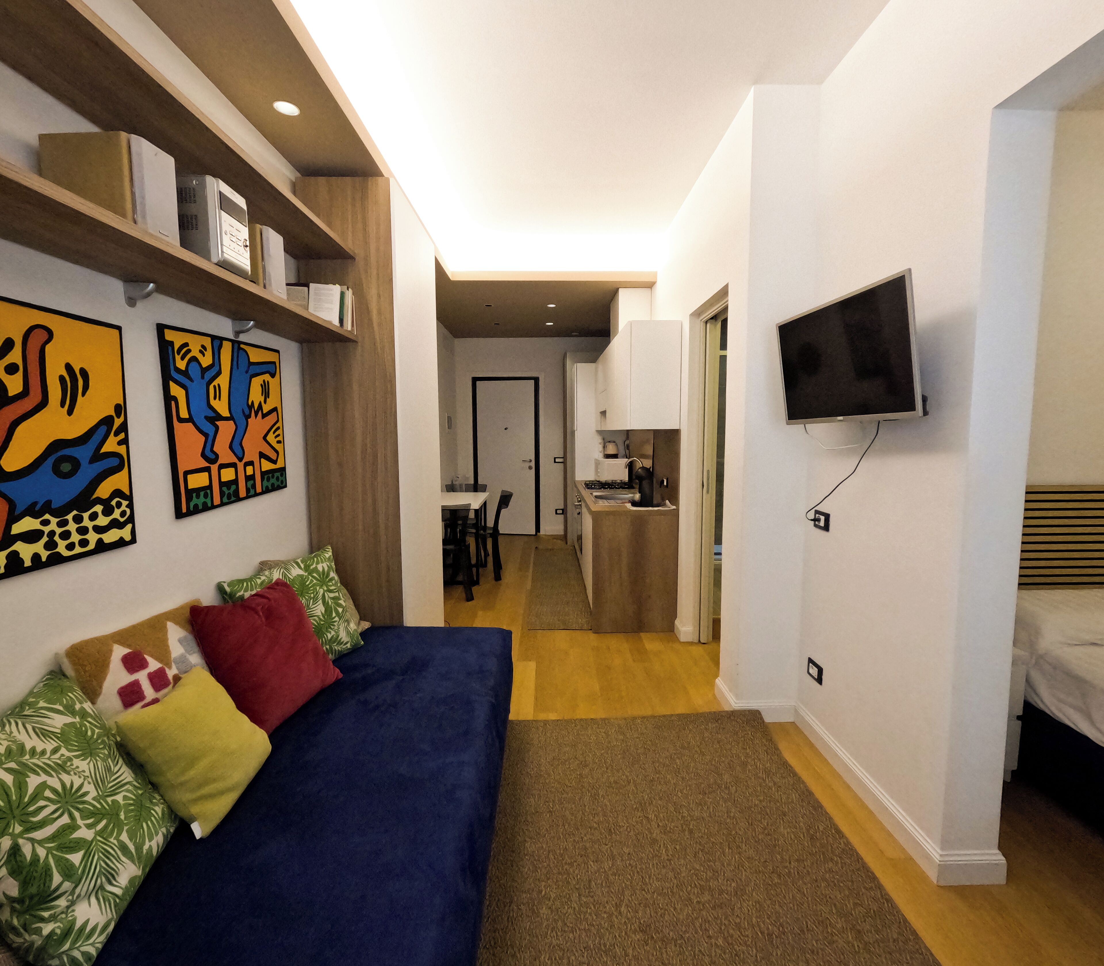 A compact living space with a sofa, a TV mounted on the wall, and a kitchenette in the background.