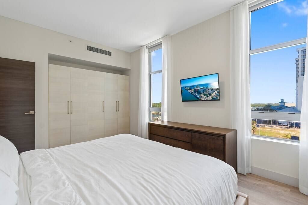 Ein Schlafzimmer mit einem großen Bett, einem an der Wand montierten Fernseher und Blick auf eine Stadtlandschaft durch ein Fenster.