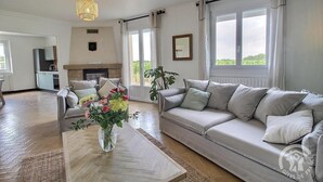 A spacious living room with a grey sofa, a coffee table with a vase of flowers, a fireplace, and large windows.