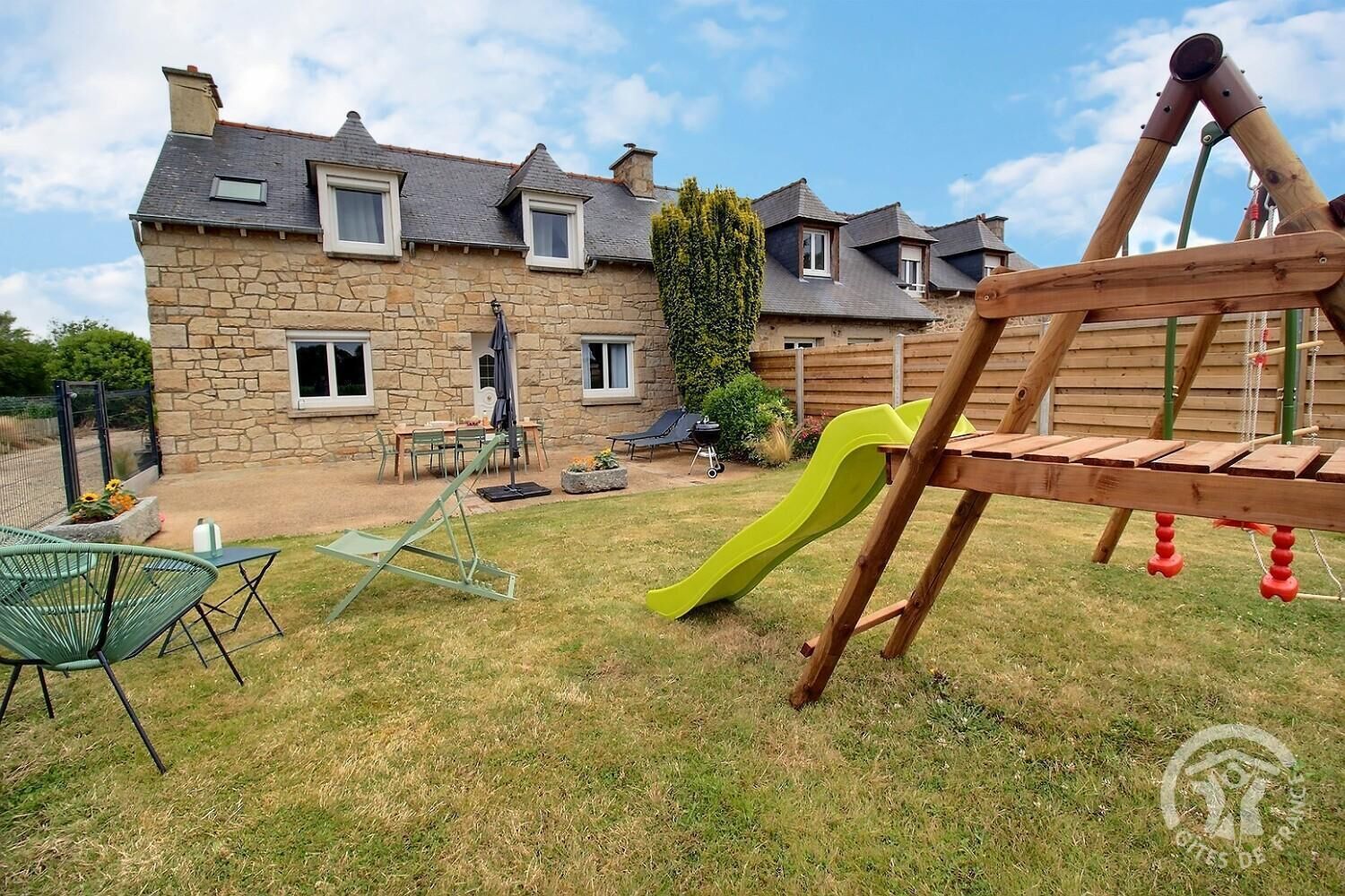A stone house with a patio, a wooden swing set, and a green slide.