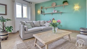 A living room with a sofa, a coffee table, a potted plant, and a wall-mounted shelf with books and a clock.