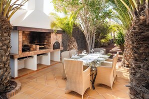 Une terrasse extérieure avec un barbecue en briques, des meubles en osier et une table en verre, le tout disposé pour les repas.