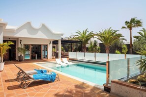 Un espace piscine extérieur moderne, doté de chaises longues, d’un patio fermé par des baies vitrées et d’une piscine entourée de palmiers et de cactus.