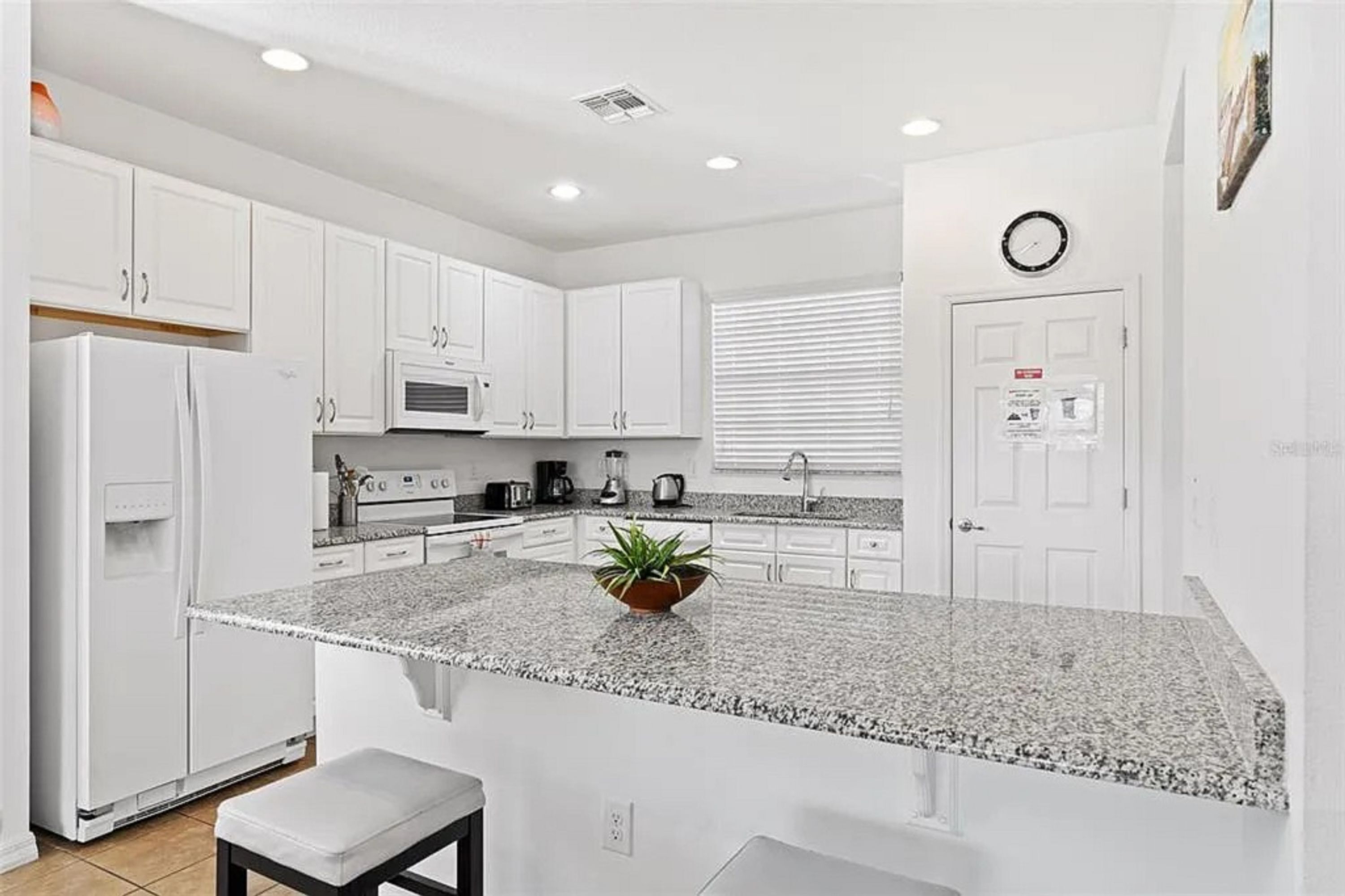 A modern kitchen with white cabinets, granite countertops, a built-in microwave, and a small island with a plant.