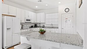 A modern kitchen with white cabinets, granite countertops, a built-in microwave, and a small island with a plant. - Grand Executive Deluxe 4BR Pool Home Near Disney Parks (Davenport)
